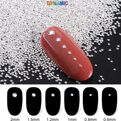 A stunning nail art display features a glossy, reddish-brown nail embellished with Dynamic Nail Supply's Metallic Mini Beads for Nail Art Designs. These beads are meticulously arranged from large to small, adding a touch of elegance with shimmering beads scattered around. The silhouettes below showcase sizes ranging from 2mm to 0.6mm, highlighting the exquisite precision achievable with this product, available in 6 grids and 6 sizes per pack.