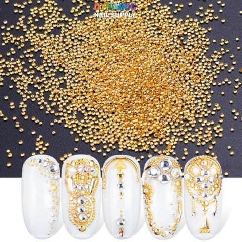 The image displays a selection of Dynamic Nail Supply's metallic mini beads, available in six different sizes per pack. Five white artificial nails are featured below, adorned with elaborate nail art that incorporates gold beads and silver rhinestones, creating intricate patterns. The composition stands out against a flat black background.