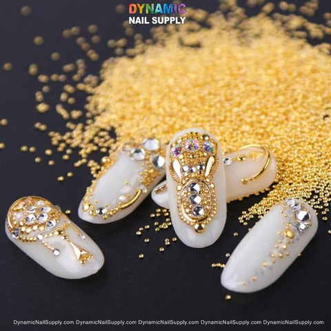 An intricately designed set of artificial nails is displayed on a black background, featuring exquisite nail art. The white nails are adorned with ornate gold decorations, such as beads, gems, and filigree patterns. Scattered in the background are metallic mini beads from the Dynamic Nail Supply's "Metallic Mini Beads for Nail Art Designs - 6 grids / 6 sizes / per pack," which enhance the luxurious and elaborate nail designs.