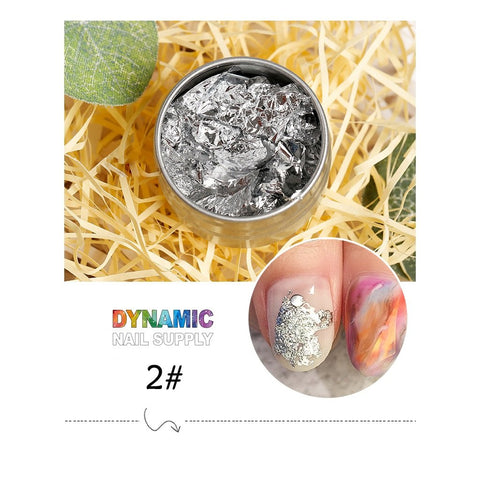 A small metal container filled with Big Size 14cm Nail Foil paper for Nail Art Design rests on a bed of straw-like material. Below, two nail designs highlight silver foil accents and a vibrant marbled pattern, crafted using Dynamic Nail Supply products. The text reads "Dynamic Nail Supply" with a colorful logo and "2#" beneath it.