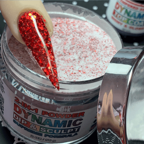 A close-up captures a hand adorned with a long, red glittery nail as it dips into an open jar of Red Holographic glitter acrylic powder - 366 - KE078 by Dynamic Nail Supply. The powder within is a pale pink interspersed with red specks, and the silver lid rests nearby. In the background, a blurred container of Dynamic Top Coat suggests endless possibilities for nail designs.