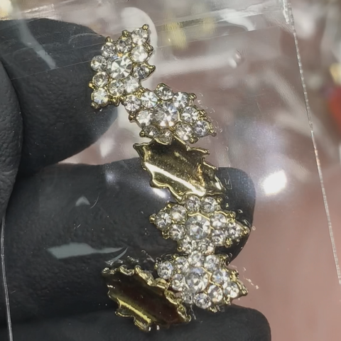 A close-up photo highlights a rhombus-shaped charm held by a gloved hand, from Dynamic Nail Supply's 6-piece set designed for Nails Art Design. The golden-base accessory boasts a zigzag pattern adorned with clusters of clear rhinestones and leaf-like elements. The blurred background accentuates the charm's intricate design and sparkle.