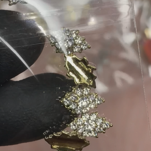 A gloved hand holds a piece of gold jewelry adorned with clusters of small, sparkling diamonds, much like the intricate design created by the 6 pcs Rhombus shape Charm for Nails Art Design (Golden base) by Dynamic Nail Supply. Captured against a reflective surface, its brilliance is enhanced by the clear gemstones set closely together.