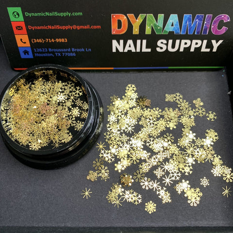A black container with scattered gold snowflakes-shaped sequins is placed on a black surface. Behind it lies a business card displaying "Dynamic Nail Supply" in vibrant text, with contact details like a phone number and an address in Houston, TX. This setup is ideal for creating festive nail art designs using the Snowflakes sequins - Christmas Metal Sequins from Dynamic Nail Supply.