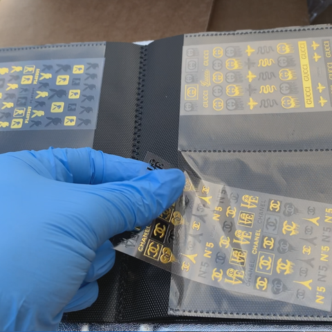 A gloved hand is carefully holding a transparent sheet of yellow and gold nail stickers against a black page from the chic Stickers Album by Dynamic Nail Supply. The sheet showcases various luxury brand logos, such as Chanel and Gucci, with 120 stickers neatly arranged in rows. This elegant collection is elegantly stored within the hard cover Stickers Album, which boasts 60 pages accommodating sticker sheets measuring 3.5" x 4.5".