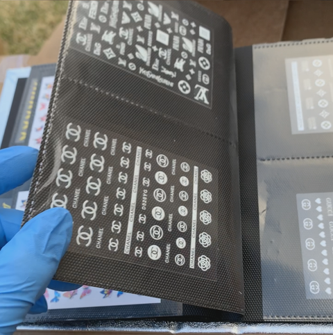 A person wearing blue gloves flips through a hardcover Stickers Album by Dynamic Nail Supply, which contains 60 pages and holds 120 white nail art decals organized on transparent sheets. The black folder features various designs, including logos and geometric shapes, all neatly displayed for easy viewing.