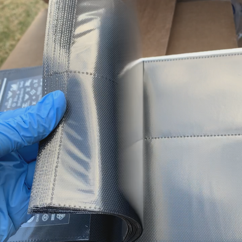 A gloved hand holds an open roll of shiny black liner material, featuring a textured grid and visible seam lines reminiscent of the Stickers Album (hard cover) by Dynamic Nail Supply. In the background, a cardboard box can be seen.