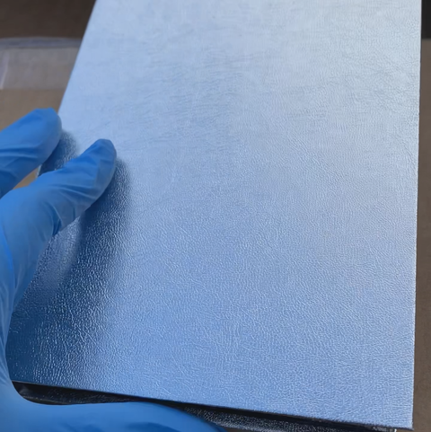 A gloved hand carefully touches the silver, textured hardcover of the Stickers Album by Dynamic Nail Supply, its metallic sheen reflecting light. The blue glove implies gentle handling as it emerges from a cardboard box, suggesting an inspection. This tactile experience could belong to this exquisite album in pristine condition, capable of holding 120 stickers sized 3.5" x 4.5" across its 60 pages.