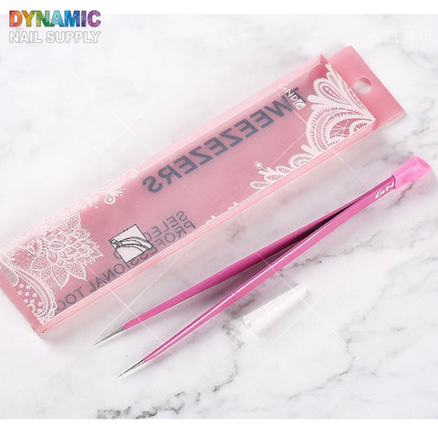 The pink pointed nails art tweezer with a cap rests on a marble surface next to its transparent packaging, adorned with white lace designs and the Dynamic Nail Supply branding.