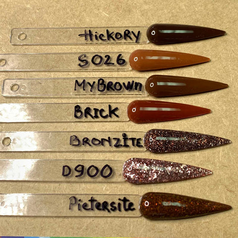 Six nail polish swatches on transparent sticks are labeled: Hickory (light brown), 5026 (medium brown), MyBrown (dark brown), Brick (reddish-brown), Bronzite with glitter from nail powder, D900 featuring gold shimmer from acrylic powder, and Pietersite highlighting a dark brown fine shimmer. These shades are available in the Dynamic 2-in-1 Powder - Nude Collection 05 - Part 2 by Dynamic Nail Supply.