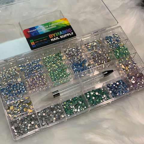 An open organizer box made of clear plastic, holding the [OPAL IRIDESCENT MULTI COLOR] Rhinestones set by Dynamic Nail Supply, is displayed on a fluffy white surface. This set includes rhinestones in vibrant iridescent colors with various shapes and sizes—14 big shapes and 6 round shapes arranged in 20 cells. A precision wax pen for easily picking up these dazzling beauties accompanies the set. A card featuring the Dynamic Nail Supply brand and contact information is also prominently visible.