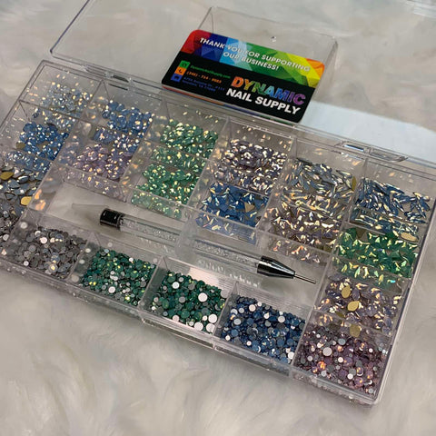 A [OPAL IRIDESCENT MULTI COLOR] Rhinestones set from Dynamic Nail Supply, featuring 20 compartments with vibrant iridescent rhinestones in shades of blue, green, pink, and gold, is organized neatly in a clear plastic case. In the center rests a nail art tool alongside a Dynamic Nail Supply card. All these items are displayed on a fluffy white surface.