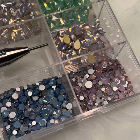 A close-up of the [OPAL IRIDESCENT MULTI COLOR] rhinestones set by Dynamic Nail Supply shows a plastic container with 20 compartments, each holding assorted iridescent rhinestones. The sections are filled with teardrop-shaped pieces in blue, green, purple, and gold hues. Beside the container rests a metal-tipped nail supply tool on a soft white surface.