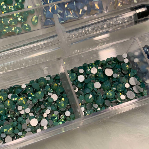 A close-up of a clear plastic container from Dynamic Nail Supply, showcasing their [OPAL IRIDESCENT MULTI COLOR] Rhinestones set. The container is divided into 20 compartments, each filled with round and uniquely shaped rhinestones. The main section features an assortment of shiny green and silver gems, while the background compartment displays iridescent light blue gems. A jewel picker rests on top.