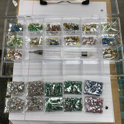 An open Dynamic Nail Supply [OPAL IRIDESCENT MULTI COLOR] Rhinestones set, featuring 20 compartments filled with assorted small embellishments like iridescent rhinestones in shades of green, blue, and silver, sits atop a lined notebook. A clear plastic pen is placed beside this dazzling nail supply collection.