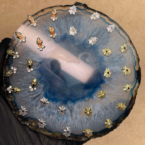 A hand, wearing a black glove, holds a polished geode slice with a blue center. On the geode rest Halloween Nails 3D Charms by Dynamic Nail Supply—including stud earrings and festive charms like bats, ghosts, spiders, and spiderwebs in silver and gold hues. The arrangement perfectly sets the scene for imaginative Halloween nail art against a neutral beige backdrop.