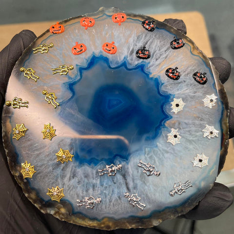A gloved hand showcases a blue agate slab adorned with Halloween Nails 3D Charms from Dynamic Nail Supply, ideal for nail supply enthusiasts. The charms are arranged in a circular pattern and feature vibrant orange and black jack-o-lanterns, skeletons, and spider webs against the vivid blue center of the agate.