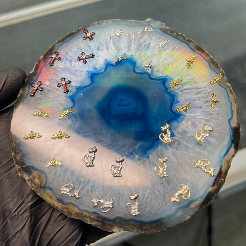 A gloved hand holds a polished agate slice featuring a vibrant blue center, embellished with Dynamic Nail Supply's Halloween Nails 3D Charms in the form of cats and crosses, some sparkling with rhinestones. Ideal for crafting unique Halloween nail art, the agate displays bands that transition from blue to white and pale gray.