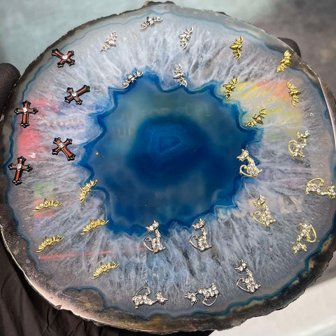 A gloved hand displays a circular blue agate with a crystalline center, complemented by stud earrings in the shapes of silver and gold crosses, anchors, and leaves. The intricate patterns of the agate reflect shades of blue, white, and gray—creating an ideal base for nail art enhanced with Halloween Nails 3D Charms from Dynamic Nail Supply.