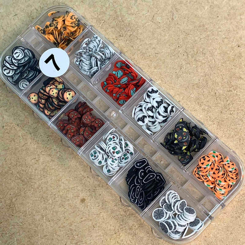 A clear plastic organizer from Dynamic Nail Supply's Halloween Nails Glitter Sequins for Nail Art Designs - Fall Nails 2021 collection contains a variety of colorful, small clay slices and a hint of cosmetic glitter in different designs. Each compartment showcases unique patterns, including animal faces, emoji-like expressions, and abstract designs. The case is placed on a wooden surface with a white sticker labeled 1.