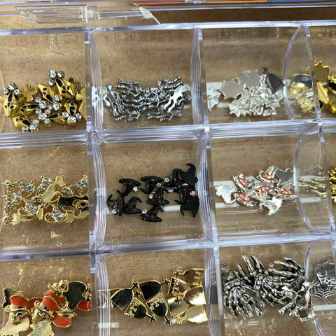 Discover Dynamic Nail Supply's Halloween 3D Charms Bundle Set for nail art design, neatly organized in a divided transparent box. This set includes assorted decorative charms in gold, silver, and black hues with various shapes such as crowns, stars, and bows. Some 3D nail charms are embellished with small rhinestones or colored accents. The box is displayed on a wooden surface.