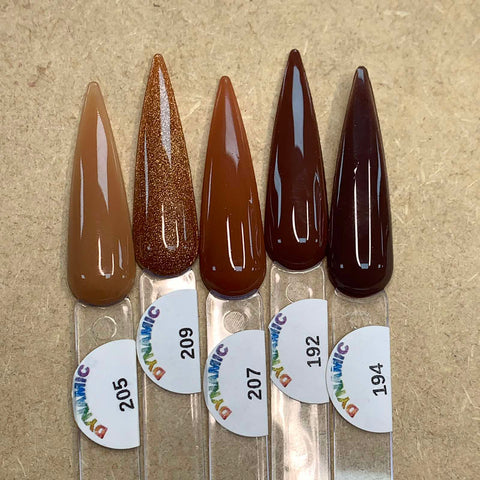 Five nail polish samples from the Dynamic Nail Supply's 2024 New Fall Colors Acrylic Collection (Part 1) - Brown Shades for Fall Season are artfully displayed on a textured surface. The colors span from light beige (205), shimmering bronze (209), medium brown (207), dark copper (192), to deep chocolate brown (194). Each chip-resistant shade is labeled with a number on a clear plastic stick, making them perfect for nail art.