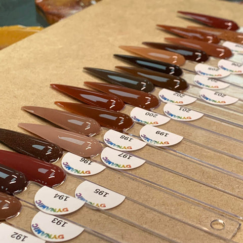 Displayed on a beige surface are samples from the 2024 New Fall Colors Acrylic Collection (Part 1) by Dynamic Nail Supply. Each chip-resistant nail polish color is presented on a clear, elongated stick with color code labels featuring shades from light nude to dark brown, each proudly bearing the Dynamic brand name in colorful letters.