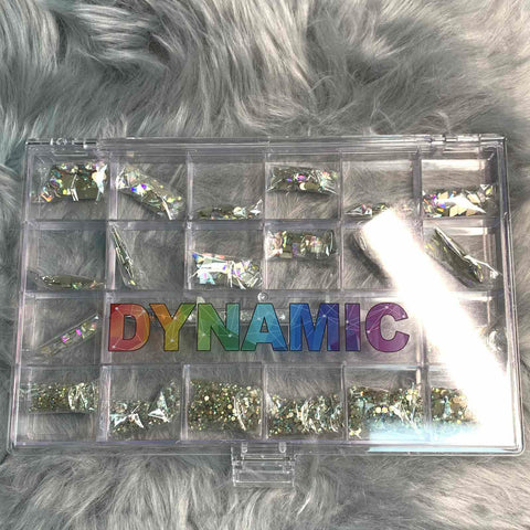 A clear plastic organizer with 20 compartments containing various AB K9 crystal rhinestones, including 14 large shapes and 6 round shapes from Dynamic Nail Supply. The word DYNAMIC in rainbow colors is printed across the lid. The background features a fluffy, gray fur-like fabric, perfect for using a rhinestone picker.