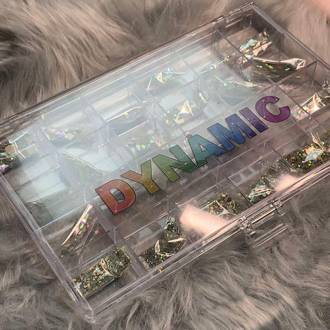 A clear plastic organizer with holographic trim rests on a soft, grey faux fur surface. Inside, the AB K9 Crystal Rhinestone set from Dynamic Nail Supply is displayed, featuring iridescent foil fragments and a rhinestone picker. The word DYNAMIC is printed on the lid in rainbow gradient letters, evoking the vibrant style of Dynamic Nail Supply.
