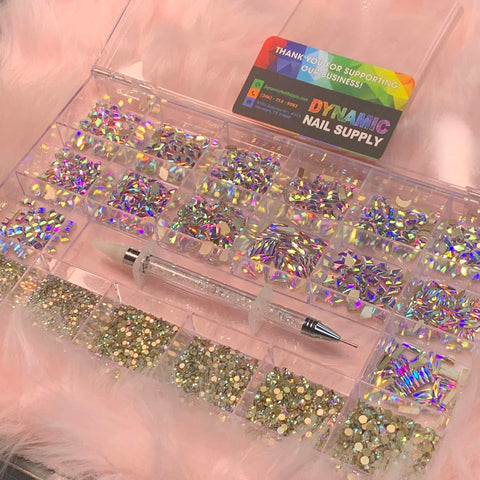 A clear organizer containing the AB K9 Crystal Rhinestone set from Dynamic Nail Supply, featuring 14 big shapes and 6 round shapes of vibrant rhinestones, is displayed on a soft pink surface. A rhinestone picker sits in front of the open box, alongside a Dynamic Nail Supply business card expressing gratitude to customers for their support.