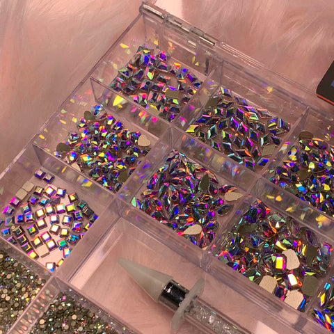 A container from Dynamic Nail Supply, equipped with 20 compartments, holds a variety of AB K9 Crystal Rhinestones in different shapes and sizes, including 14 big shapes and 6 round shapes. Nearby, there is a rhinestone picker with a pointed tip for easy use. The background features soft pink plush fabric.
