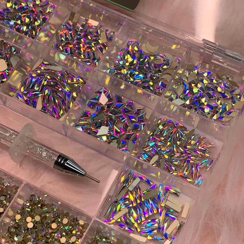 A clear organizer features the AB K9 Crystal Rhinestone set from Dynamic Nail Supply, containing an assortment of iridescent rhinestones in 14 big shapes like triangles and rectangles, along with 6 round shapes that reflect vibrant rainbow colors. Nearby, a precision-tip rhinestone picker from Dynamic Nail Supply rests on a soft pink surface, ready for use in nail art design.