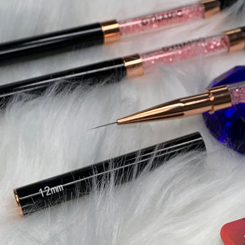 Close-up of three Dynamic Nail Supply Dynamic® Professional Dual-head Liner Brushes with black handles and pink glitter-filled sections, arranged diagonally on a white, fluffy surface. A brush cap labeled 12 mm is next to one brush, exposing its precise tip. A dark blue, faceted object is partially visible in the scene.