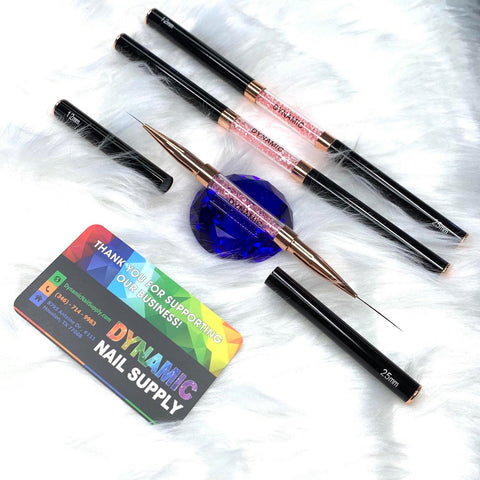 On a soft white fur backdrop, the sophisticated arrangement of nail art tools showcases three Dynamic® Professional Dual-head Liner Brushes, each with sleek black handles and metallic accents encircling a blue gemstone. A Dynamic Nail Supply card is prominently displayed in the foreground, featuring their social media and contact information, along with two brush caps nearby.