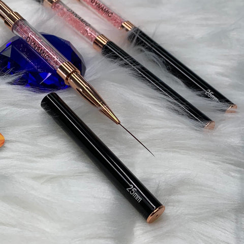 A close-up of three Dynamic® Professional Dual-head Liner Brushes for fine lines showcases their sleek black handles and rose gold accents, elegantly resting on a fluffy white surface. Each brush is marked with 25mm near the base, while one uncapped brush reveals a thin, precise bristle tip. A partially visible blue gem in the background enhances the essence of Dynamic Nail Supply.