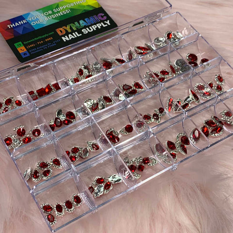 On a soft pink surface lies a clear plastic organizer box showcasing the 120 pcs Red Nail Charms set with Red Gems for nail art design and decoration. A business card from Dynamic Nail Supply is elegantly placed at the top left corner of the box.