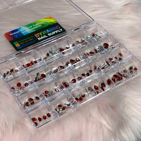 A clear plastic box rests on a fluffy pink surface, featuring multiple compartments filled with shiny red and silver nail art decorations, including the dazzling pieces from the 120 pcs Red Nail Charms set with Red Gems for nails art design and decoration. Inside the box lid is a colorful thank you card from Dynamic Nail Supply.