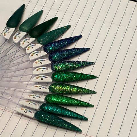 A fan of nail polish swatches, part of the Green Acrylic Collection by Dynamic Nail Supply, is displayed on lined paper. The non-toxic swatches display a range of shades from dark green to glittery emerald, each labeled with a number. The sticks are arranged in a gradient from matte greens to sparkling finishes, featuring colors like Jade, Elegant Green, Enchanted Green, Metallic Green, Kelly Green, Seaweed, Mystery Green, and Garland Green.