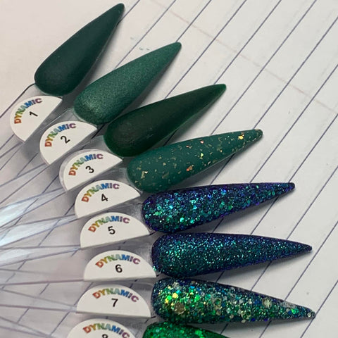 A display of nail polish samples on a notebook highlights the non-toxic Green Acrylic Collection from Dynamic Nail Supply. The collection features shades ranging from matte and glossy dark greens to sparkly greens and blues, each labeled with numbers 1 to 8. The samples are presented in an elongated, pointed shape with the word DYNAMIC in colorful letters prominently displayed.