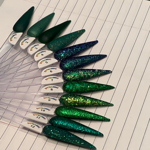 A fan-shaped nail swatch display from Dynamic Nail Supply showcases twelve long, pointed samples on lined paper. Crafted using non-toxic materials, it features the Green Acrylic Collection in a range of stunning shades from solid green to glittery tones with green, blue, and gold sparkles. Each sample is labeled with a small sticker near its base and includes colors like Jade, Elegant Green, Enchanted Green, Metallic Green, Kelly Green, Seaweed, Mystery Green, and Garland Green.