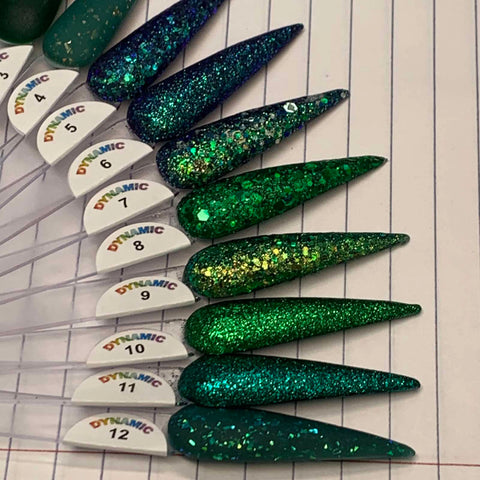 Close-up of nail polish samples on a lined surface, enhanced with acrylic nail powder. The samples display a gradient from dark blue glitter (#5) to various shades from the Green Acrylic Collection—including Jade, Elegant Green, Enchanted Green, Metallic Green, Kelly Green, Seaweed, Mystery Green, and Garland Green—ending with green glitter (#12). Each nail is marked with a number and the Dynamic Nail Supply brand name as it rests on the notebook's lined page.