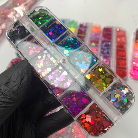 A person wearing a black glove holds a plastic container filled with Dynamic Nail Supply's "Heart Shape Holographic Glitters mixed sizes for Valentine's Day Nails Design - New 20220204-01," featuring vibrant sequins in shades of green, purple, orange, and red. These shiny, holographic glitters are perfect for creating festive nail art. In the background, additional containers with more holographic glitters add extra shimmer to the scene.