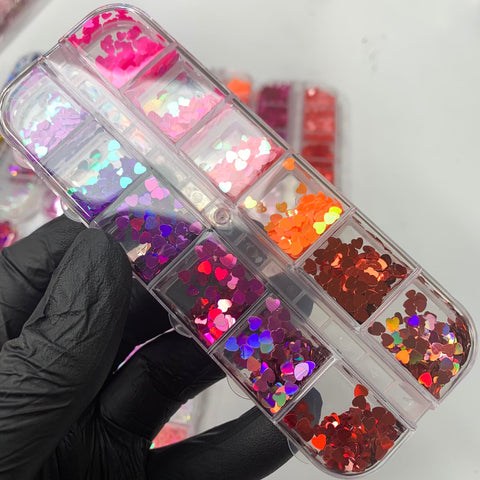 A hand in a black glove holds a transparent container from Dynamic Nail Supply, featuring their Heart Shape Holographic Glitters. This container includes twelve compartments filled with shiny and reflective, heart-shaped sequins in shades of pink, purple, red, and orange, perfect for creating vibrant Valentine's Day nail designs. In the background, similar Dynamic Nail Supply containers can be seen slightly blurred.