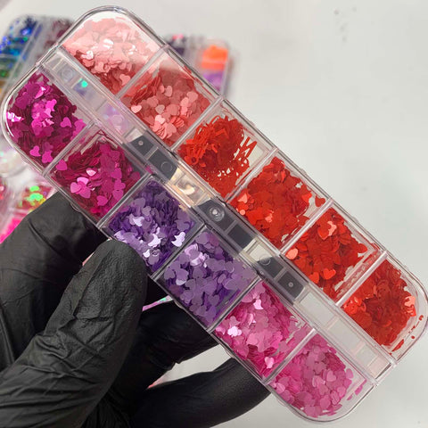 A hand wearing a black glove holds a clear plastic container filled with the Heart Shape Holographic Glitters from Dynamic Nail Supply, ideal for Valentine's Day nail designs. Each compartment glistens with captivating matte colors in shades of red, orange, pink, and purple. In the background, another container shines with even more heart shape holographic glitters.