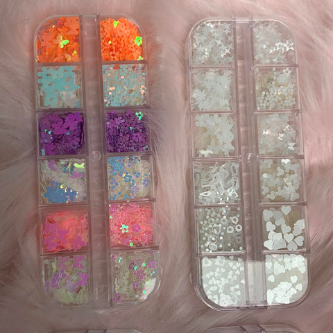 Two transparent containers from Dynamic Nail Supply, each a treasure trove of creativity, are filled with various decorative nail art glitter pieces. The left container holds vibrant sequins in delightful shapes such as butterflies and flowers, while the right features white and iridescent glitter adorned with stars and hearts. Both containers are elegantly placed on a soft pink surface.