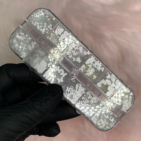 A hand wearing a black glove holds a clear plastic case displaying Dynamic Nail Supply's Heart, Butterfly, and Flower sequins in white for Spring Nails Art design. The background is set against a soft and fluffy pink surface.