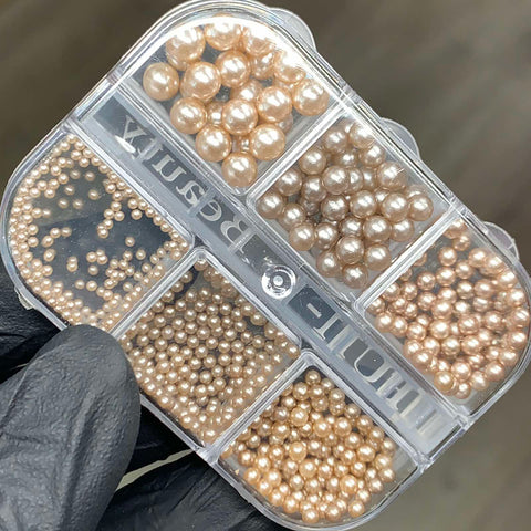 A gloved hand holds a clear plastic container with six compartments, each filled with various sizes of round, golden beads reminiscent of elegant pearls. The container is labeled "Iridescent Pearl & Metallic Ball Bead for Nails Design" from Dynamic Nail Supply. The beads have a lustrous finish and are neatly organized by size against a blurred gray background.