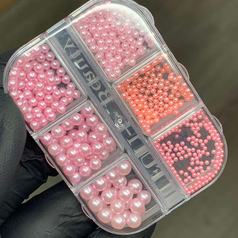 A hand in a black glove holds a rectangular plastic container featuring the Dynamic Nail Supply's Iridescent Pearl & Metallic Ball Bead for Nails Design, with six compartments each brimming with small pink and peach beads that evoke the elegance of delicate pearls. The clear lid highlights the intricate bead details, making it ideal for any nail designer seeking precision and creativity.