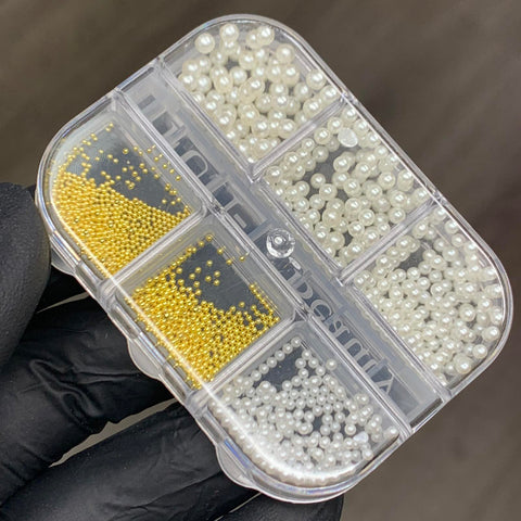 A close-up image highlights the Iridescent Pearl & Metallic Ball Bead for Nails Design from Dynamic Nail Supply, showcasing a rectangular, clear plastic container held by a gloved hand. Perfect for any nail designer, it features seven compartments filled with small iridescent white and gold beads that resemble pearls, all organized neatly against a plain, smooth gray background.