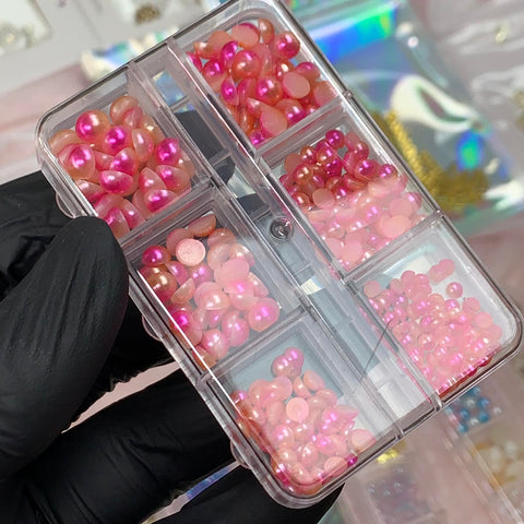A person wearing a black glove is holding a transparent plastic organizer with six compartments filled with assorted pink and red cabochons of different sizes. In the background, there's a blurred array of crafting materials and tools from Dynamic Nail Supply, ideal for use in nails design featuring their Iridescent Pearl & Metallic Ball Bead.